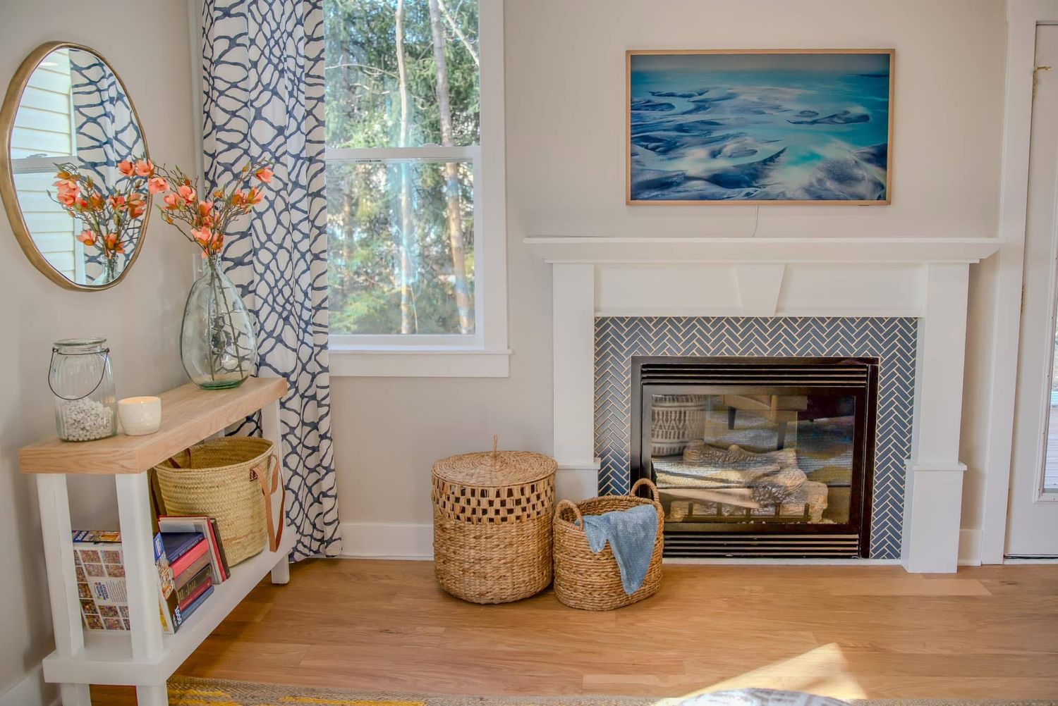 Cozy living room corner with herringbone fireplace, coastal artwork, wicker baskets, and a console table with patterned curtains.