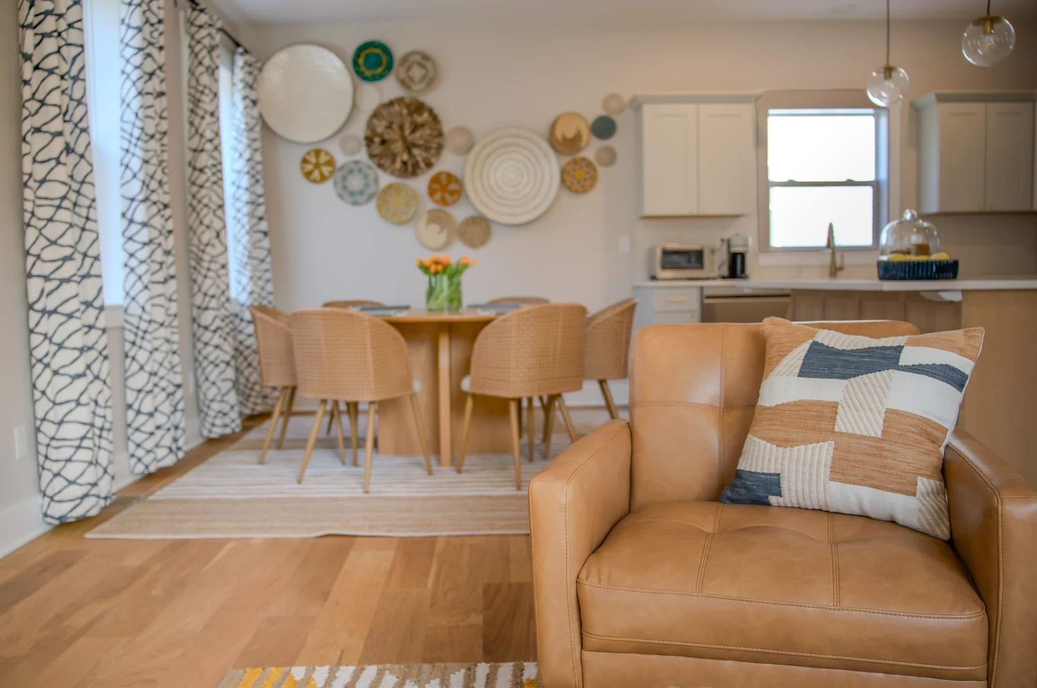 Modern living and dining area with leather armchair, round wood table, tan chairs, decorative wall plates, and open kitchen.