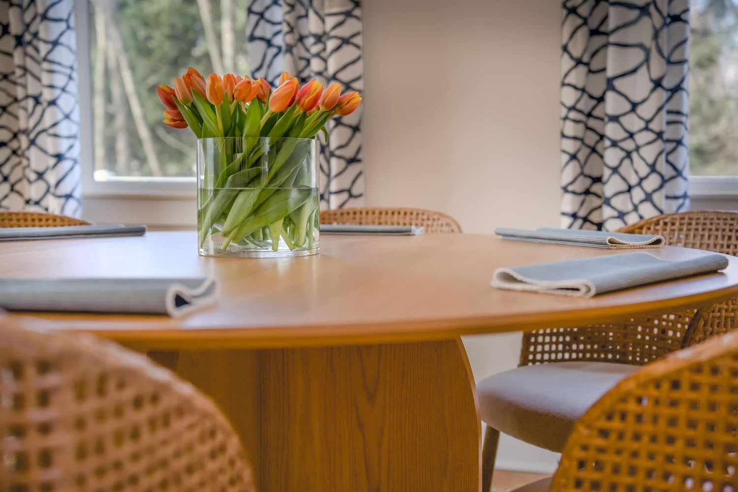 Elegant round wooden dining table with orange tulips centerpiece, wicker chairs, and patterned curtains in a bright room.