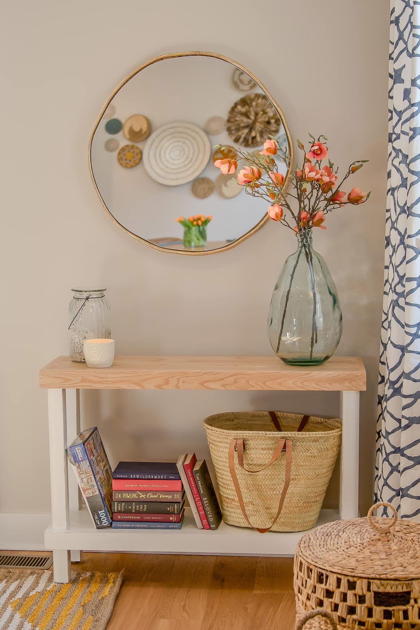 Boho entryway console table with wicker basket, stacked books, glass vase with pink flowers, and round gold mirror on beige wall.