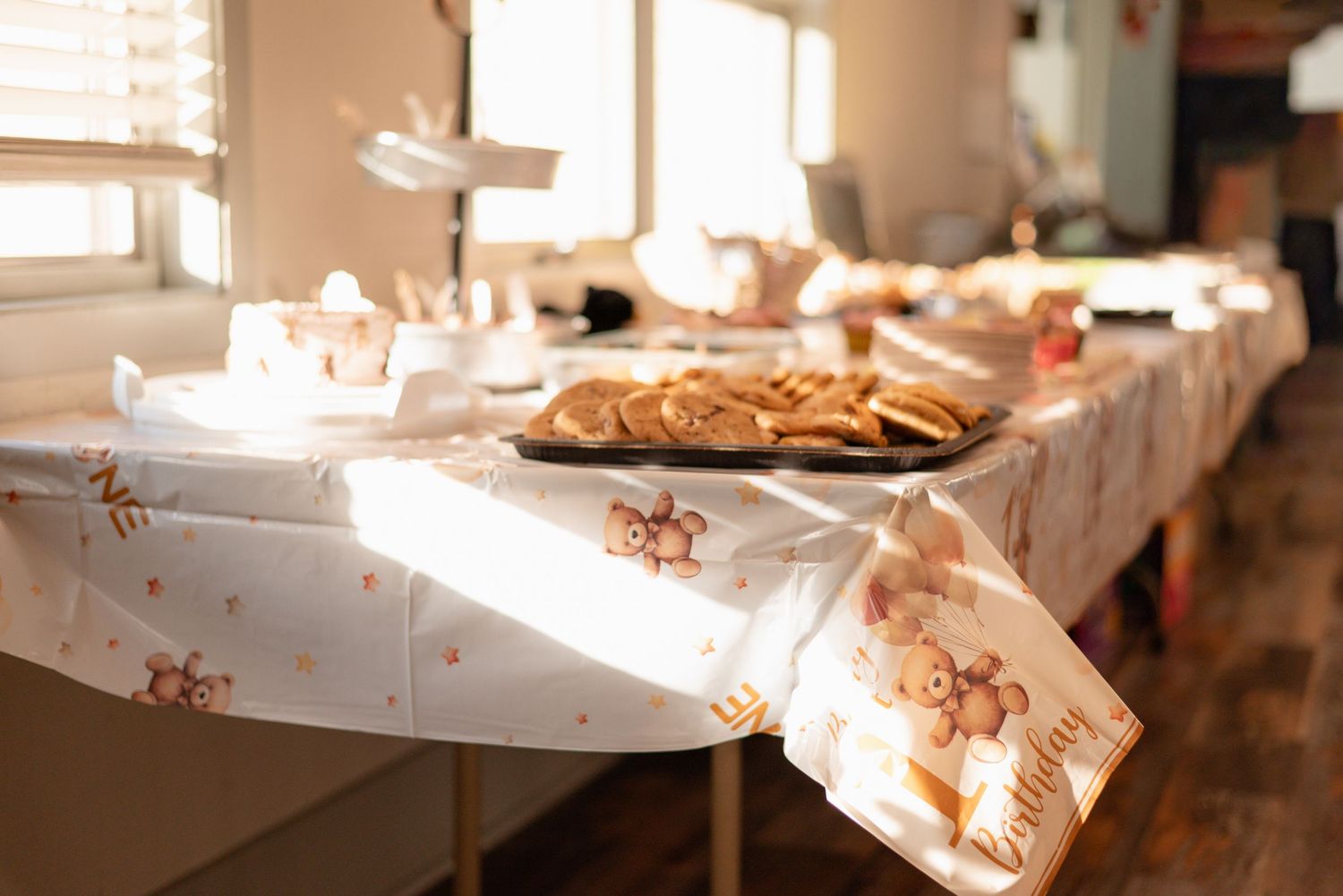 Baby shower table with teddy bear tablecloth, cookies, and treats displayed in a sunlit room.