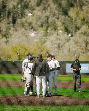 Baseball team huddles on the pitcher's mound during a game with a coach, set against a lush green hillside backdrop.