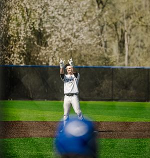 Baseball player celebrates on base with arms raised in white uniform on green field with spring trees in background.
