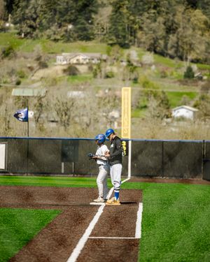 Two baseball players in blue helmets stand on base during a game with green hills and buildings in the background.