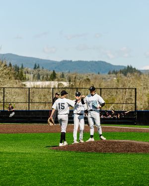 Three baseball players in white uniforms huddle on the pitcher's mound during a sunny game with green mountains behind.
