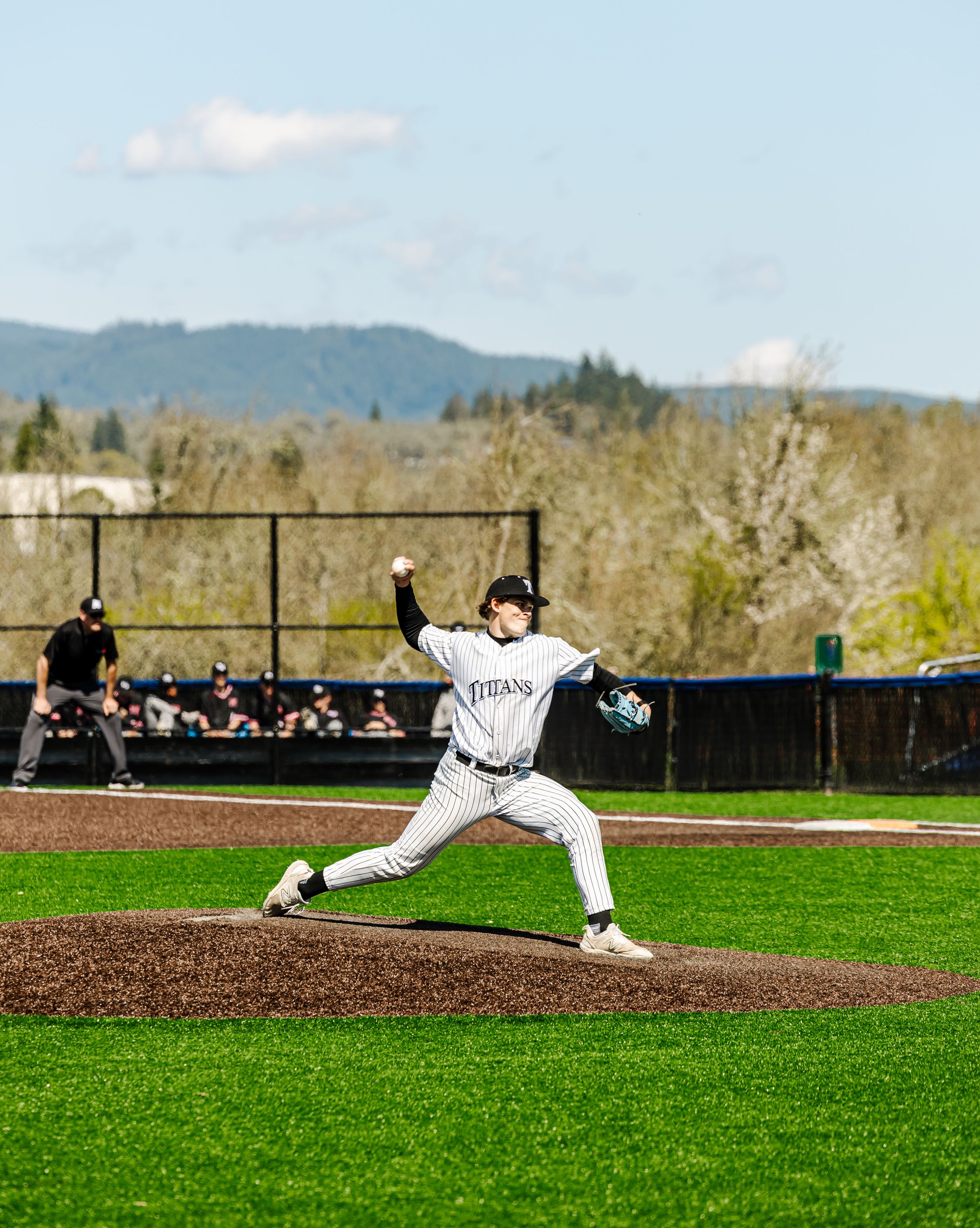 Baseball pitcher in white uniform mid-throw on a green field with trees and mountains in the background.