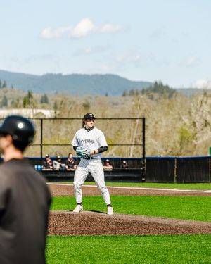 Baseball pitcher in white Titans uniform winds up on the mound during a sunny outdoor game with green hills behind.