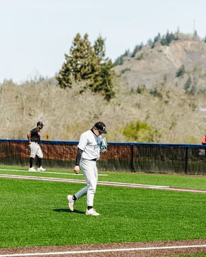 A baseball player in a white uniform walks across a green field during an outdoor game with a hill in the background.