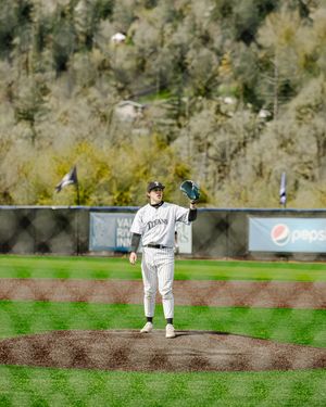 Baseball pitcher stands on mound in white uniform, glove raised, with green hillside and Pepsi sign in background.