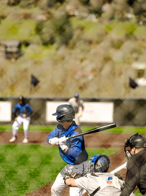 Baseball batter in blue jersey swings at pitch with catcher and umpire behind home plate during outdoor game.