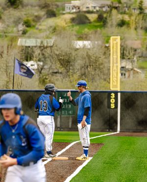 Baseball coach fist-bumps player #13 in blue uniform near third base during a spring game.