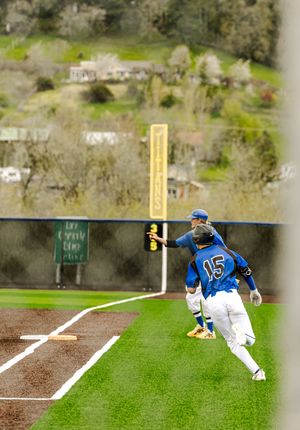 Baseball player #15 in blue uniform rounds third base during a game on a green field with tree-covered hills in background.