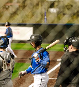 Baseball batter in blue uniform smiles at home plate with catcher and umpire during a game.