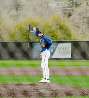 Baseball pitcher in blue jersey stands on mound, head down, preparing to pitch on a cloudy day.