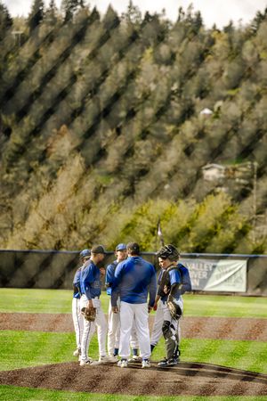 Baseball team huddles on the pitcher's mound during a game at a green field with tree-covered hills in the background.