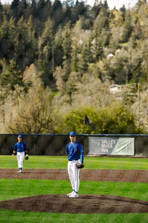 Baseball pitcher stands on mound in blue uniform at Valley River Inn field with tree-covered hills in background.