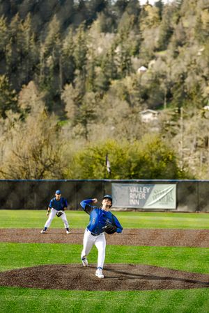 Baseball pitcher in blue uniform delivers a pitch on a green field with Valley River Inn banner and forested hills in background.