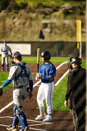 Baseball batter in blue uniform holds bat near home plate with catcher and umpire nearby.