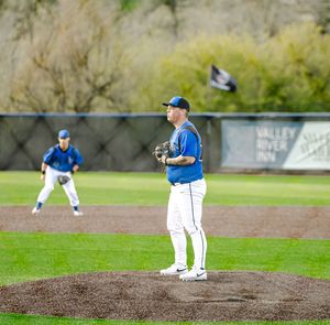 Baseball pitcher in blue and white uniform stands on mound ready to pitch with fielder positioned in background.
