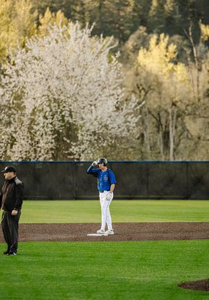 Baseball player in blue uniform stands on base during spring game with white blossoming trees in background.