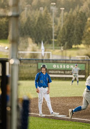 Baseball player in blue uniform standing on base during a game, with green outfield and Olsson Industrial Electric sign in background.