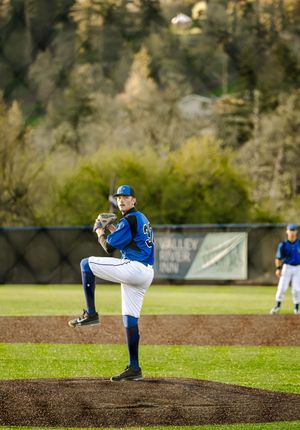 Blue-uniformed baseball pitcher winds up on the mound during a game with green trees in the background.