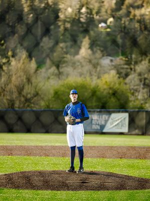 Baseball pitcher in blue uniform stands on mound ready to pitch at outdoor ballpark with green trees in background.
