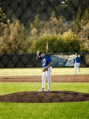 Baseball pitcher in blue uniform stands on mound, adjusting cap during a sunny outdoor game with green field.