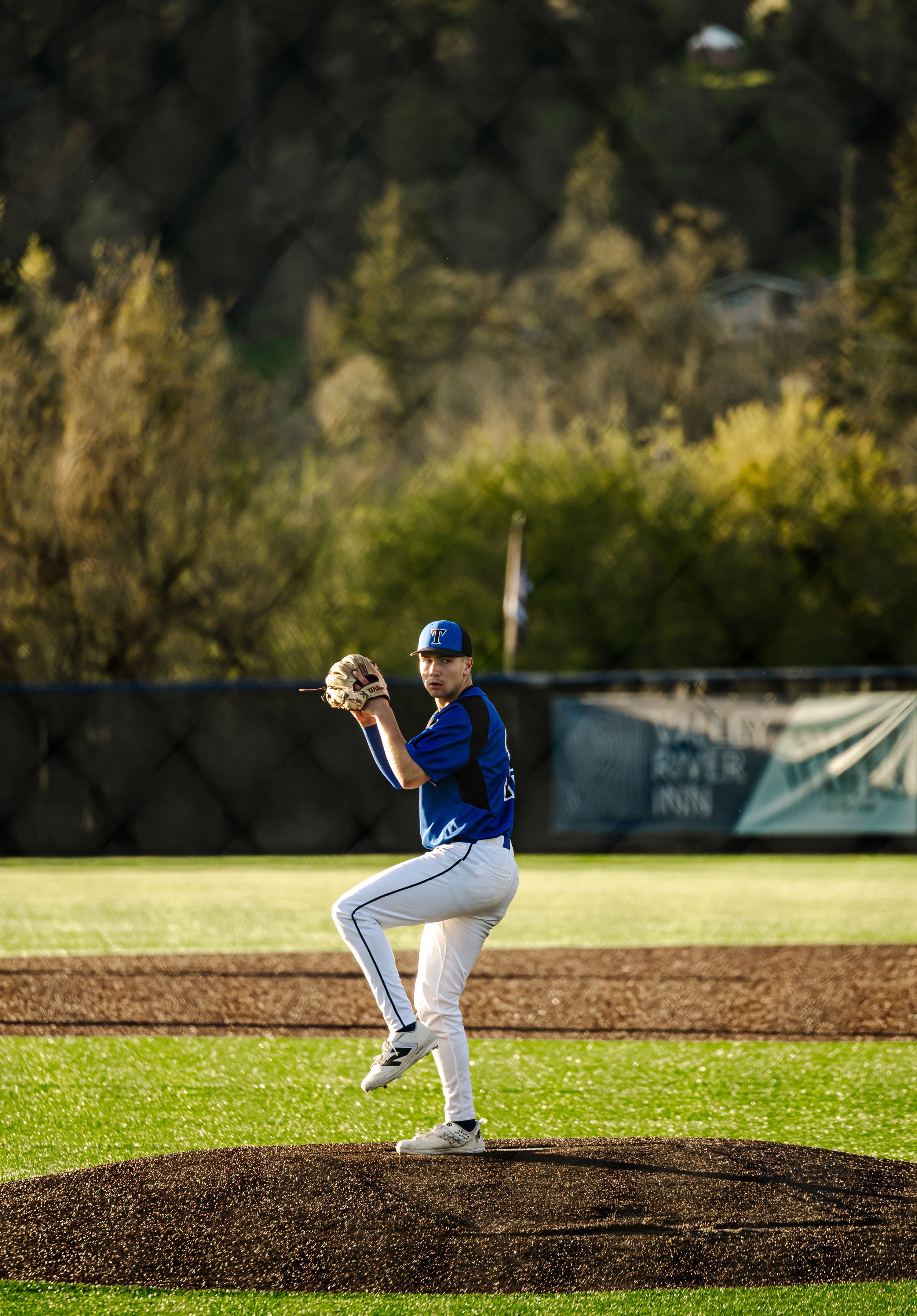 Baseball pitcher in blue uniform winds up on the mound during a sunny game with lush green trees in the background.