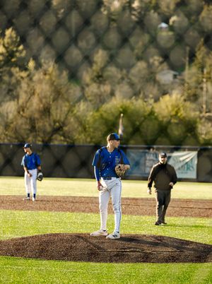 Blue team pitcher stands on mound during baseball game with teammates and umpire visible on sunny field.