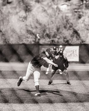 Black and white photo of baseball pitcher in mid-throw on the mound with umpire in background.