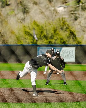 Baseball pitcher in black uniform delivers pitch on mound with umpire watching during outdoor game.