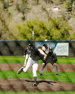 Baseball pitcher in black pinstripe uniform delivers pitch on mound during sunny outdoor game with umpire watching.