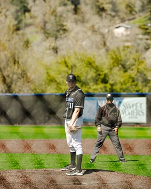 Baseball pitcher stands on mound in black uniform while umpire watches during spring game on green field.