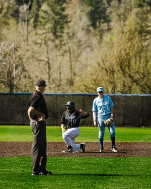 Baseball player kneels at second base as an umpire and fielder in light blue uniform look on during a game.