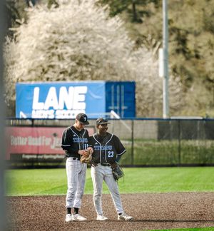 Two Lane CC baseball players in uniform confer on the field with blooming trees in the background.