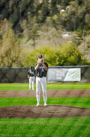 Baseball pitcher in black uniform stands on mound preparing to throw during a spring game with green hills in background.