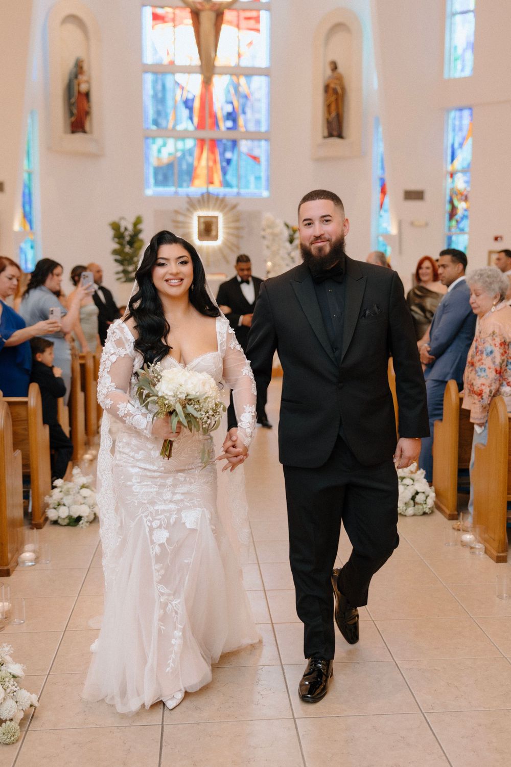 Newlyweds walk down church aisle, bride in lace gown holding bouquet, groom in black tuxedo, after ceremony.