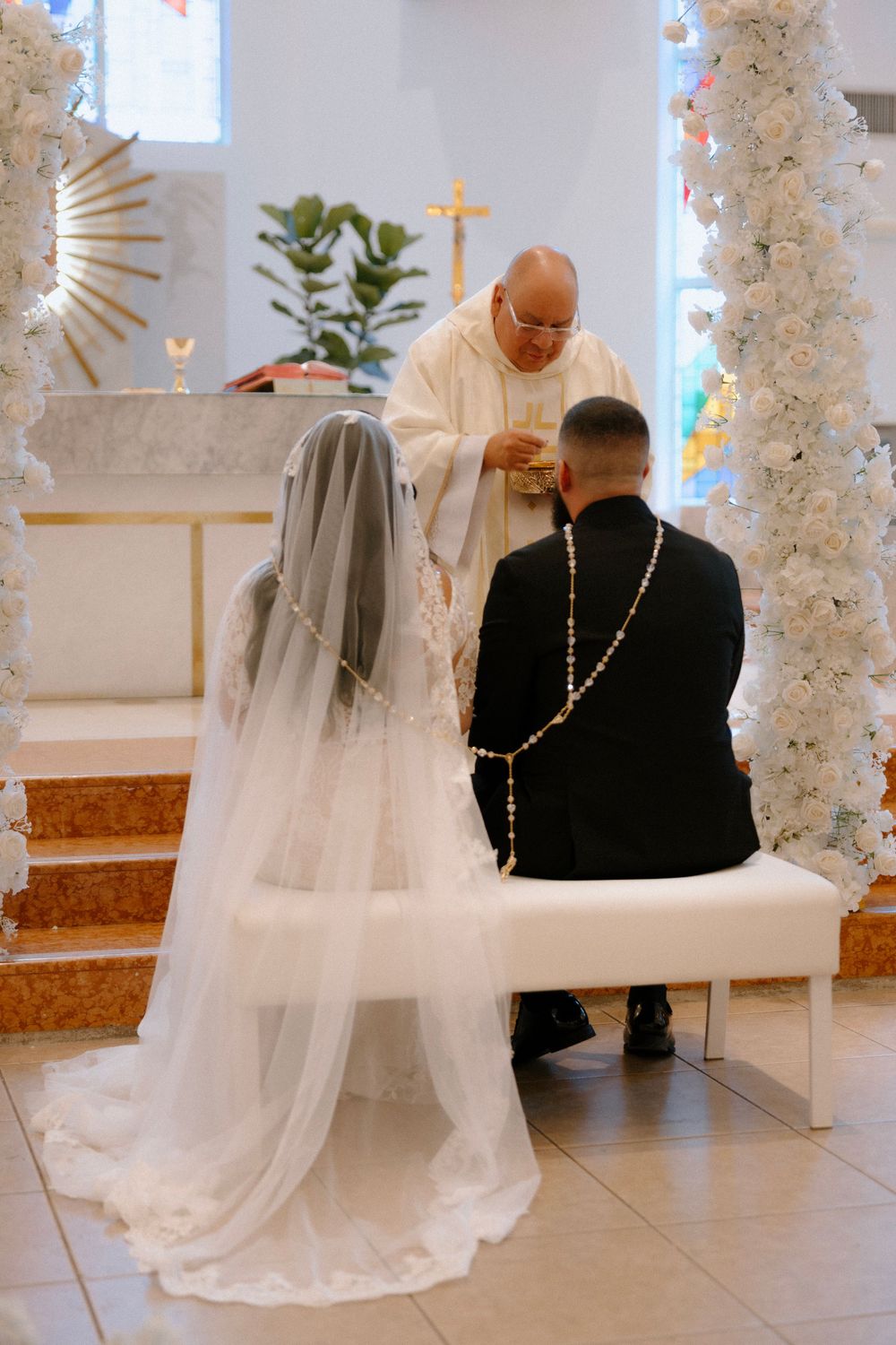 Bride and groom kneeling before priest during Catholic wedding ceremony with white floral arch decorations.