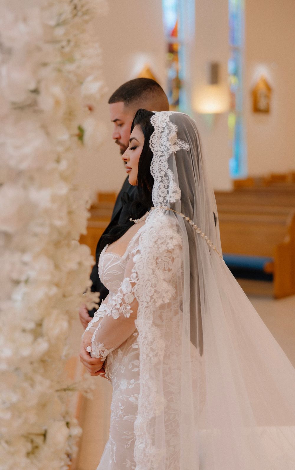 Bride in lace gown and mantilla veil stands with groom in black suit at church altar surrounded by white floral arrangements.