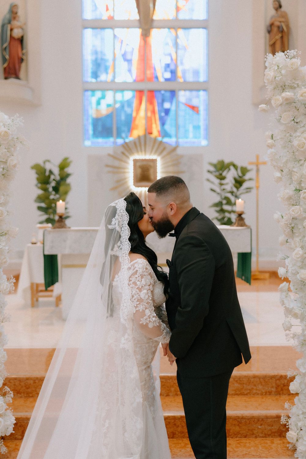 Bride and groom share first kiss at church altar surrounded by white floral arrangements and stained glass.