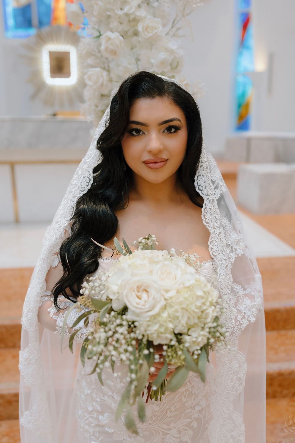 Bride in lace wedding dress and veil holding white rose bouquet, posing in elegant church setting.