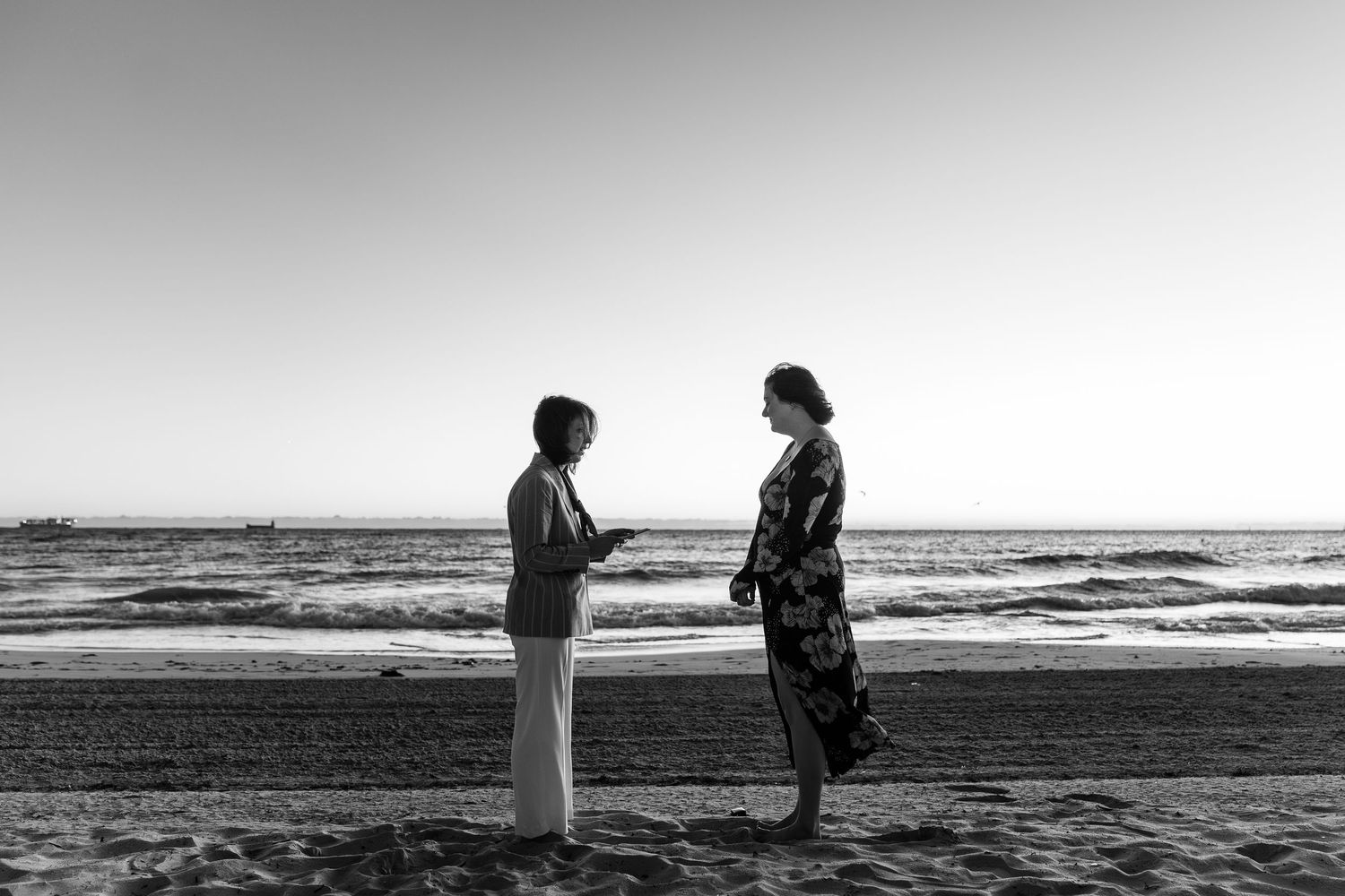 Two people stand facing each other on a beach in a black and white photo, with ocean waves in the background.