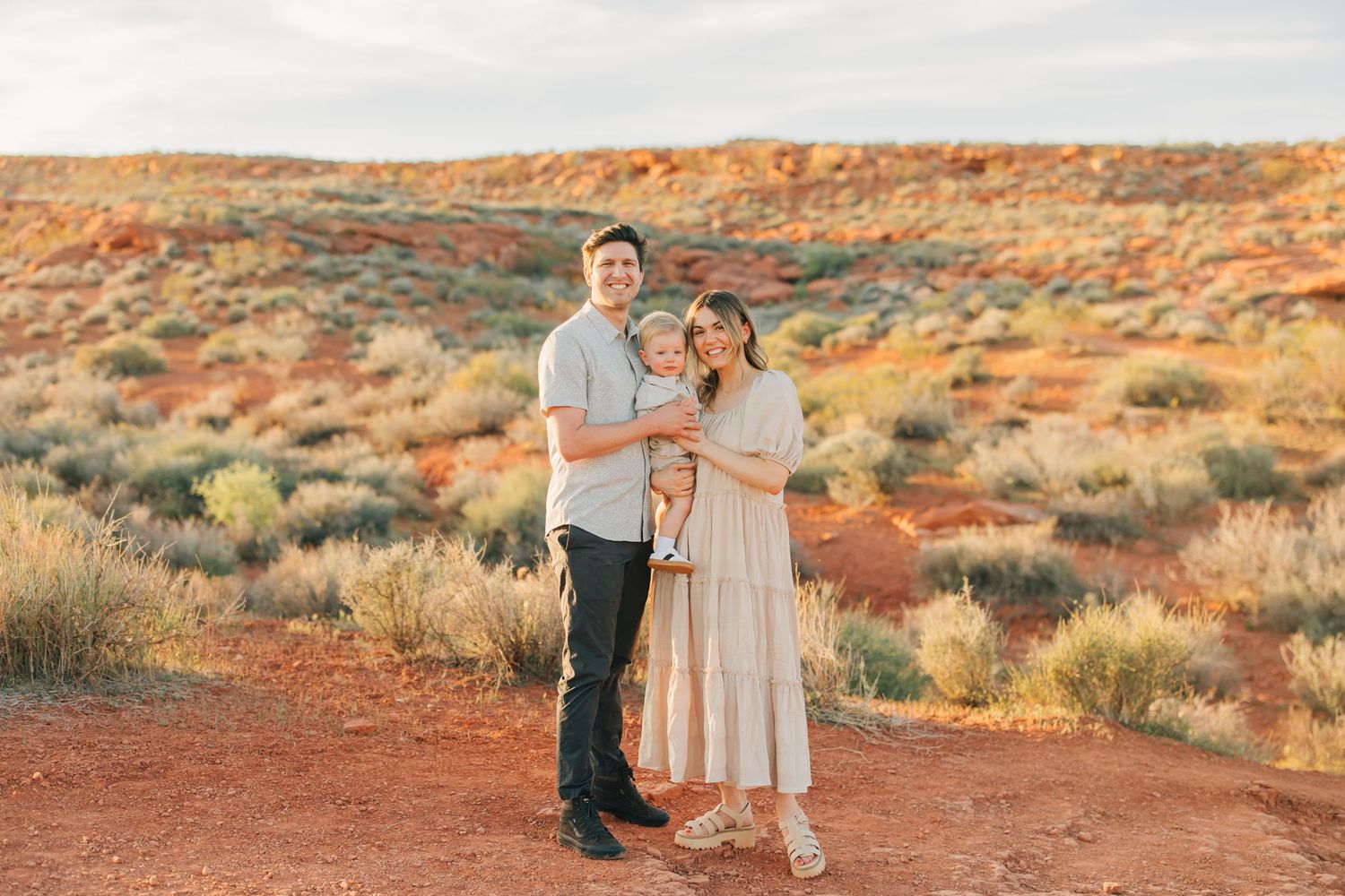 Family of three posing in a scenic desert landscape with red rock formations and golden hour lighting.