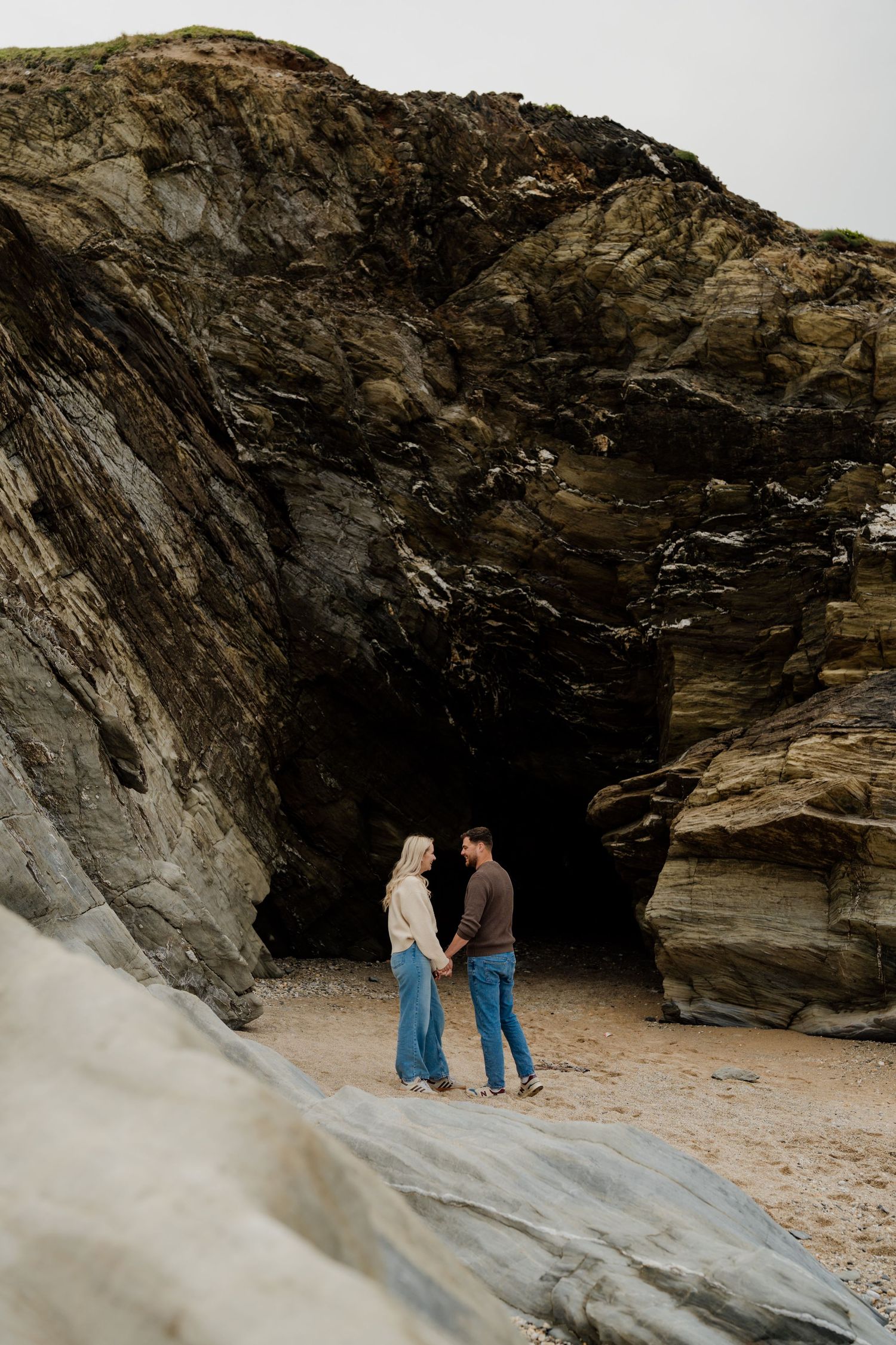 Couple holding hands on sandy beach before dramatic rocky cave cliffs on overcast day.