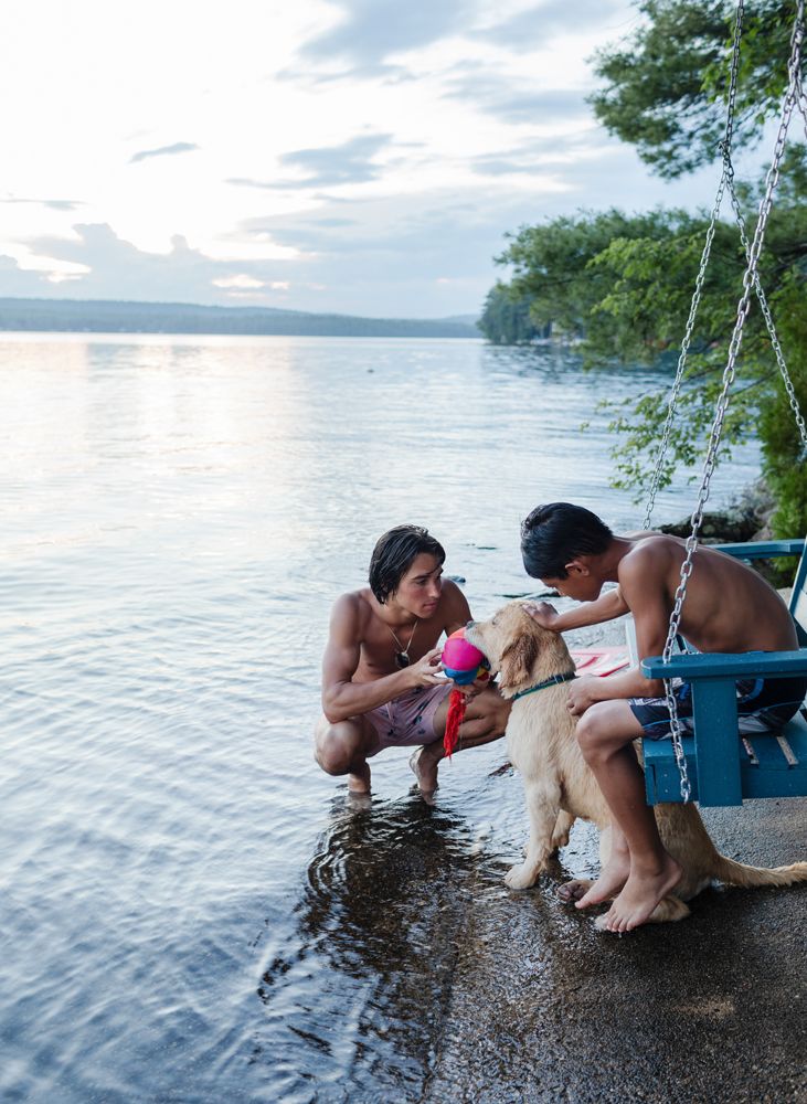 Teenage boy and younger boy pet a yellow lab crouching next to a lake on a summer evening. New Hampshire family vacation photographer.