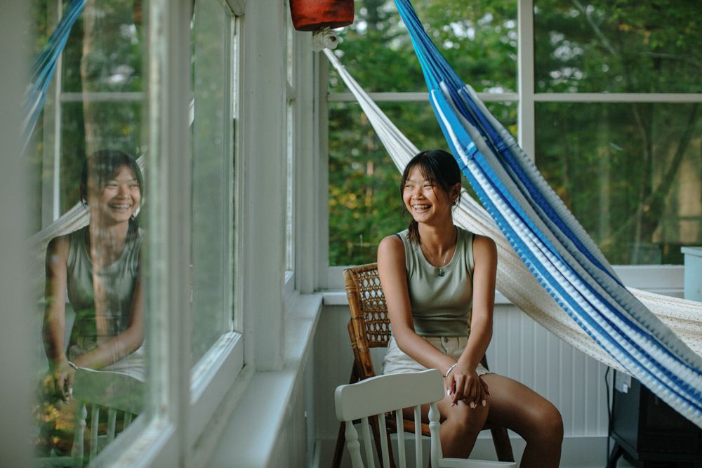 Happy girl sits on a chair near a hammock inside a cabin glass window porch with white walls, Acadia family vacation photographer.
