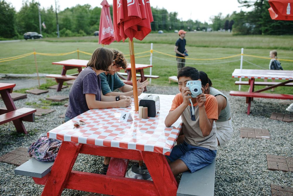 Family sits at red picnic table as boy photographs with Polaroid at a lobster pound in Acadia. Southern Maine family vacation photographer.