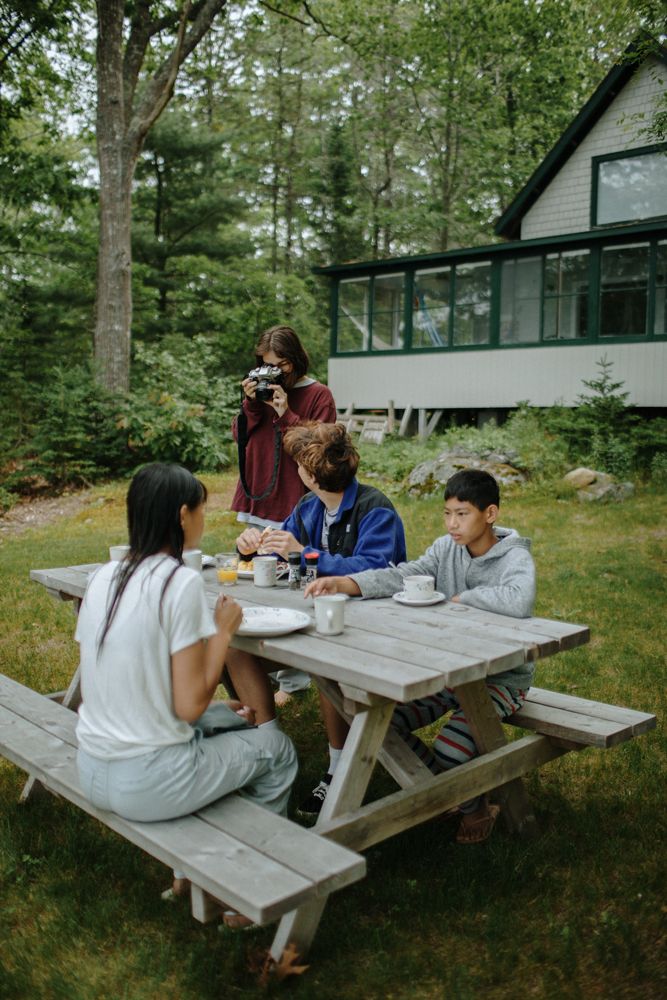 Family sits at table eating breakfast near white green cottage amidst trees as teen photographs family. Maine family vacation photographer.