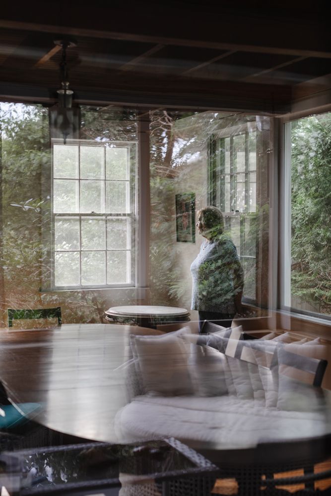 Grandmother looks at window through reflected windows in a cabin porch with a table. Cape Cod family vacation photographer.